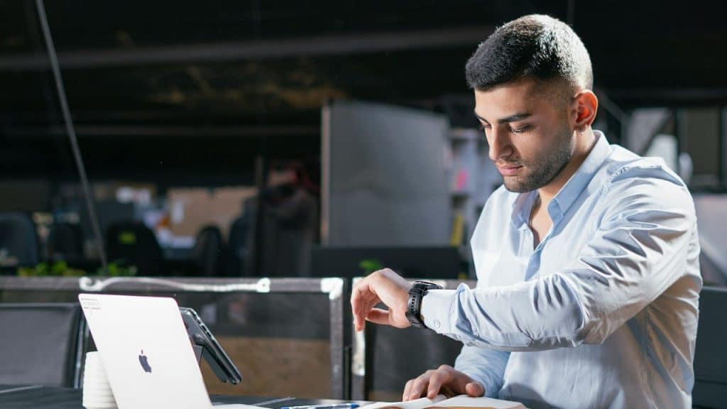 A man in a light blue shirt looks at his wristwatch while sitting at a desk with a laptop and notebook.