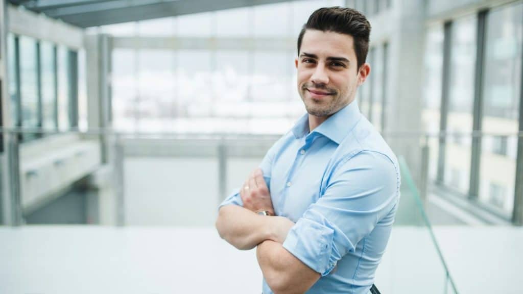 Smiling man in a blue dress shirt standing with his arms crossed in a modern office.