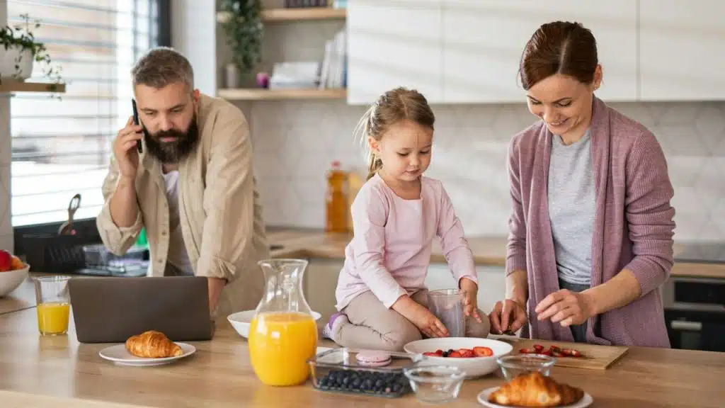 Family busy in the kitchen: man on phone, daughter on counter, woman cutting strawberries.