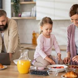 Family busy in the kitchen: man on phone, daughter on counter, woman cutting strawberries.