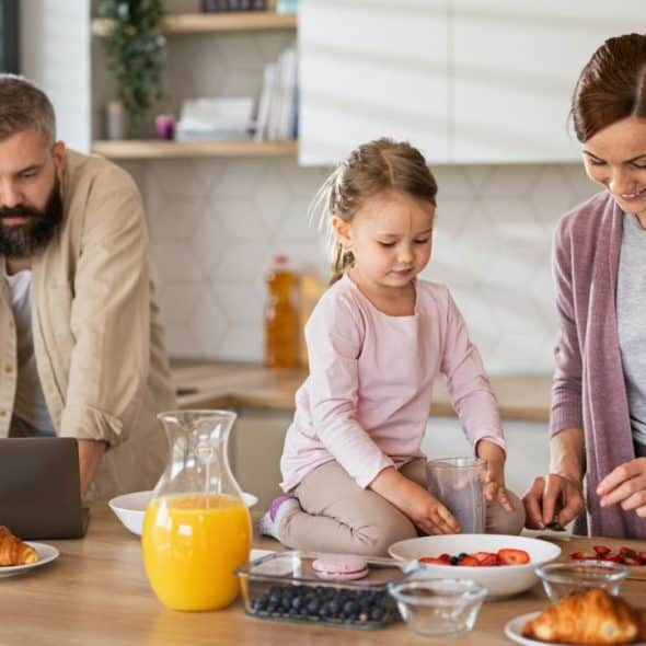 Family busy in the kitchen: man on phone, daughter on counter, woman cutting strawberries.