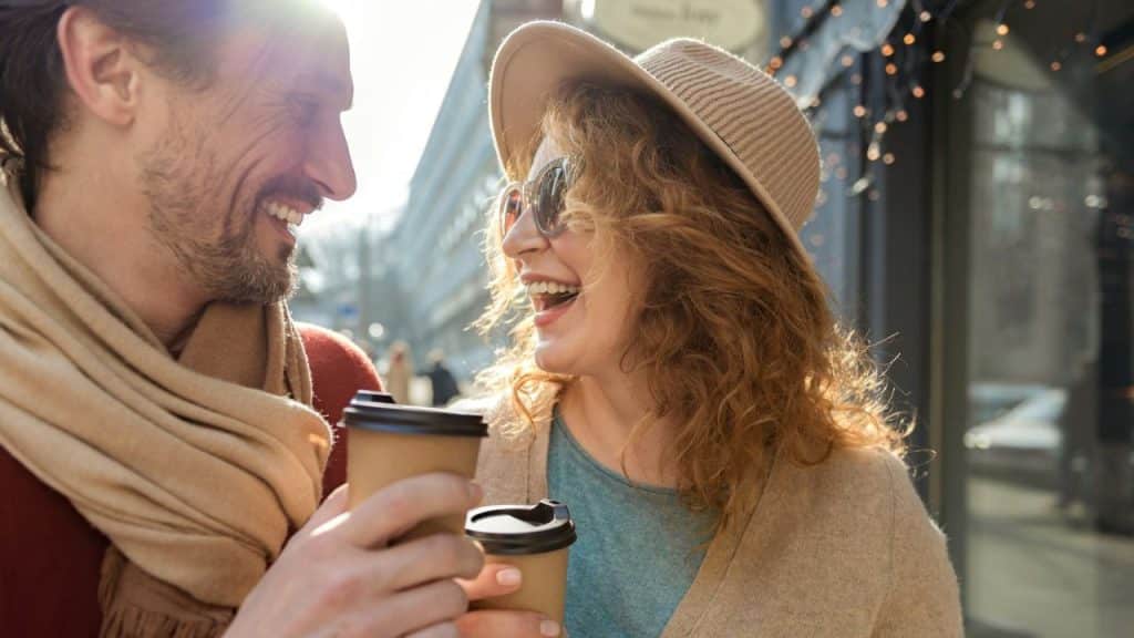 Smiling couple holding coffee cups and laughing while standing outdoors in sunlight.