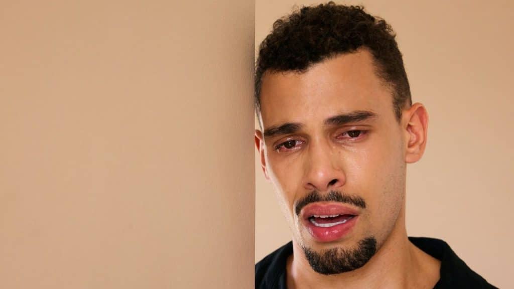 Man with teary, red eyes and a goatee crying against a tan wall.