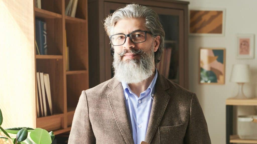 Mature man with glasses and a long white beard smiling indoors near a bookcase.