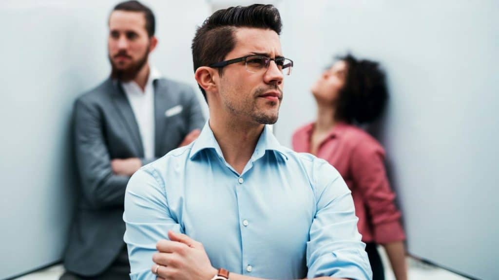 A stern man with glasses and arms crossed stands in front of two blurred colleagues.