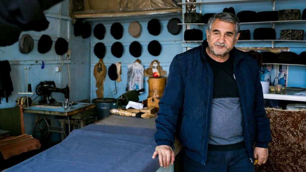 Man in a blue jacket standing in a workshop with hats on a wall.