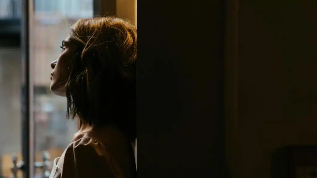 A person with short hair leaning against a wall while looking out a window.