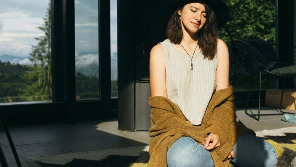 A woman sitting on the floor in sunlight, wearing a hat and sweater.