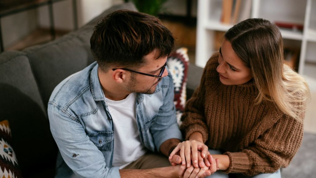 Man in a denim shirt and woman in a brown sweater holding hands and looking at each other.