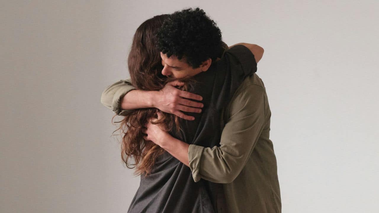 Man with curly hair hugging a woman with long hair against a plain background.