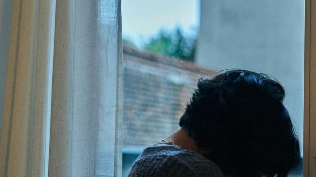A person with short dark hair sits by a window, looking outside through sheer curtains.