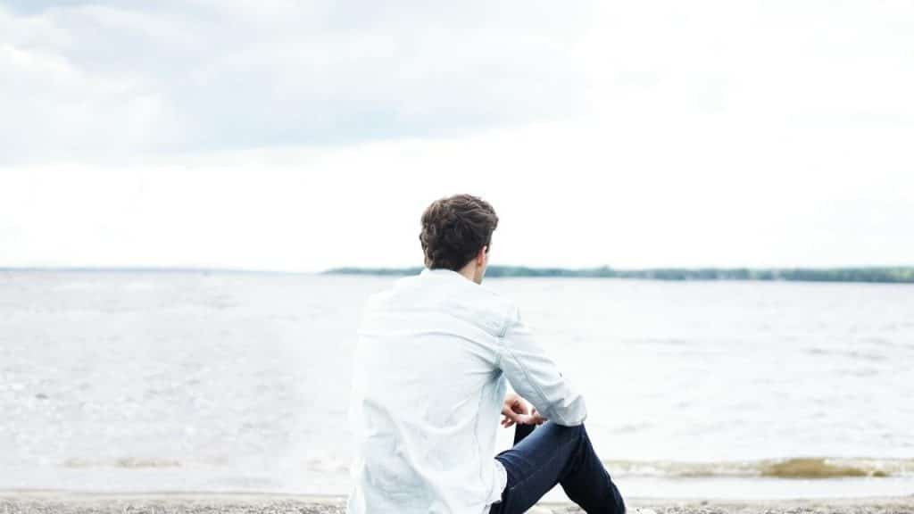 A man sitting on a beach looking out at the calm water under a cloudy sky.