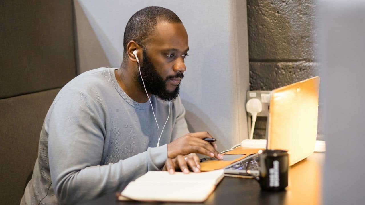 Bearded man with earbuds focusing on his laptop and notepad in a booth.