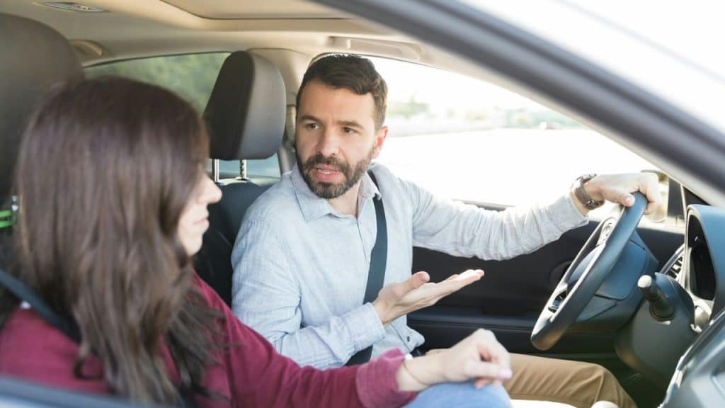 Man with a beard driving a car, talking and gesturing toward a woman passenger.