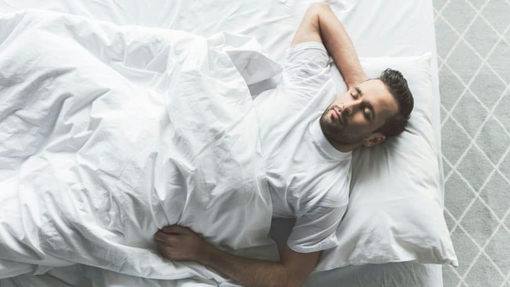 Man with a beard sleeping on his back in a bed with white bedding, high angle.