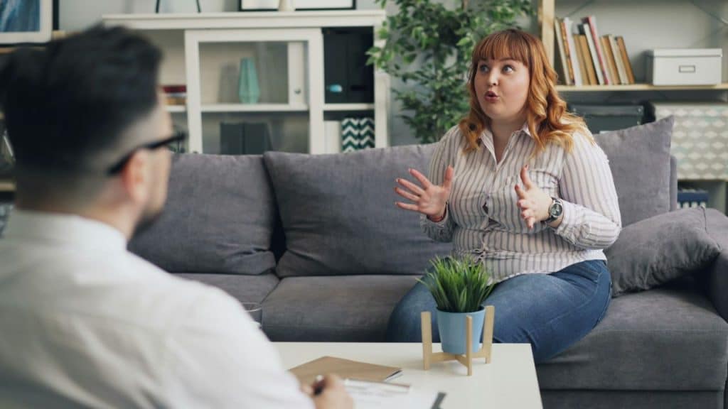 Woman sitting on a couch gesturing while speaking to a man facing away from the camera.