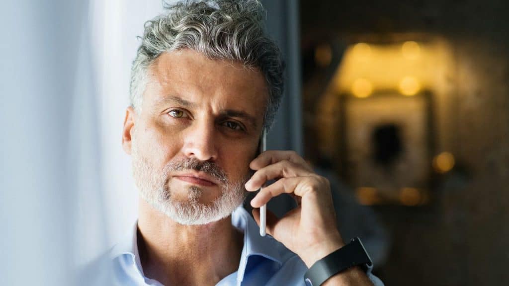 A middle-aged man with gray hair is speaking on a mobile phone, looking directly at the camera.