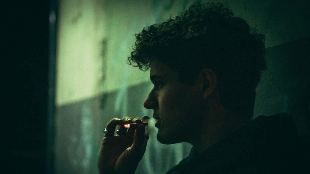 Side profile of a person with curly hair smoking a glowing cigarette in a dark setting.