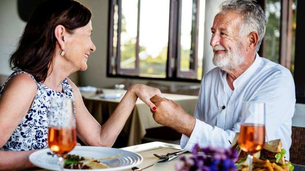 An older couple smiles and holds hands across a restaurant table with food and drinks.