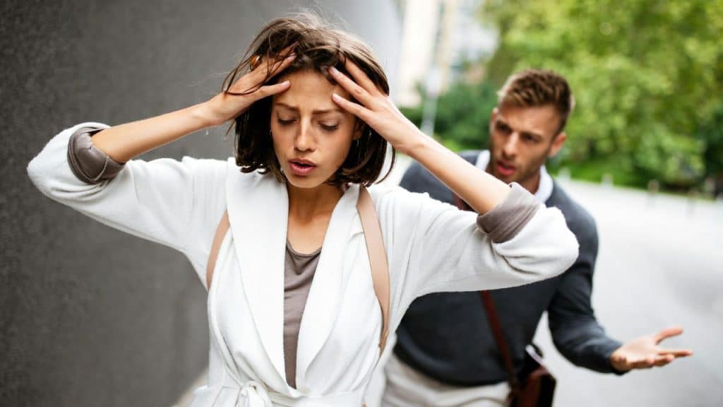A distressed woman holds her head while a man gestures in frustration behind her on a street.