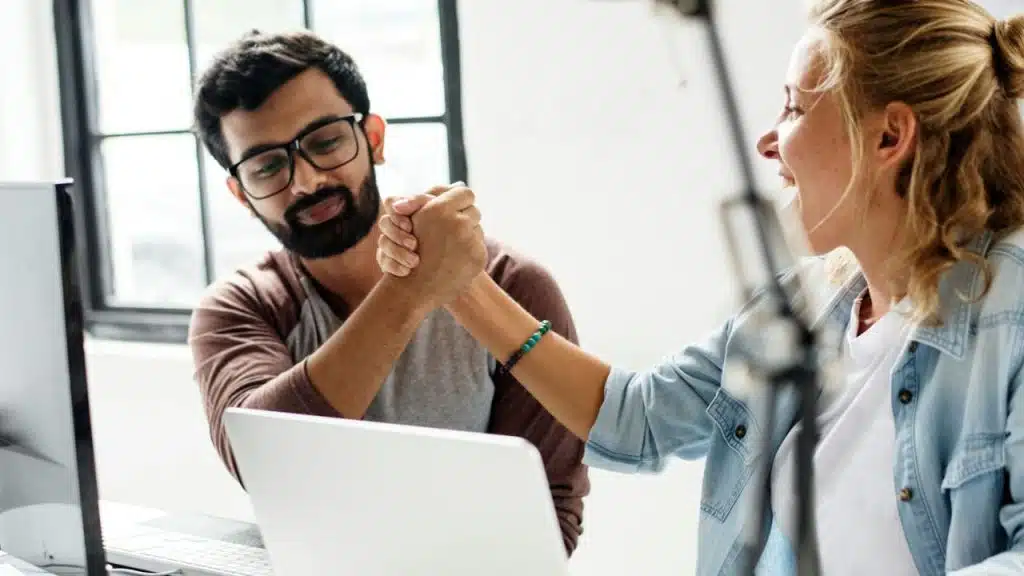 A man and a woman sitting at desks shake hands enthusiastically over a laptop.