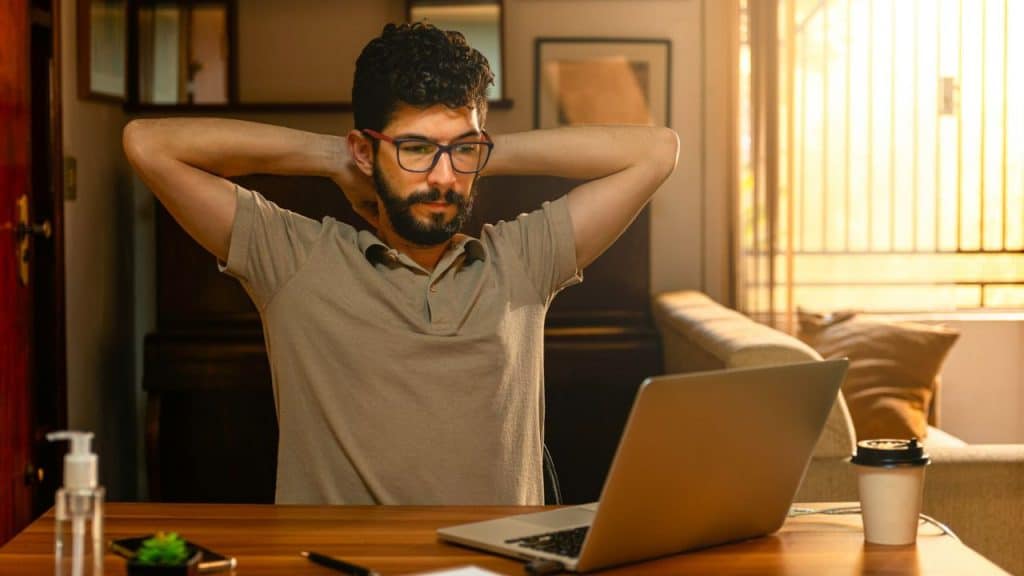 Man in glasses stretching his neck while looking at a laptop in a sunlit room.