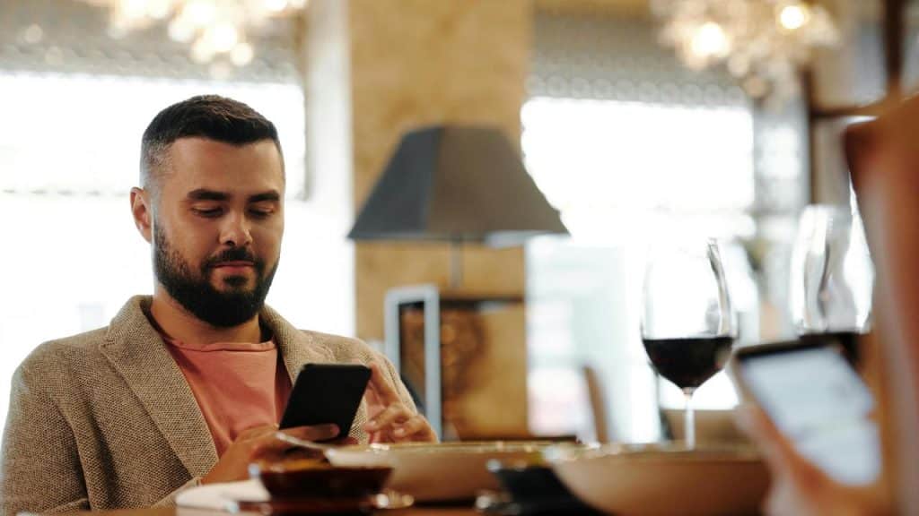 A man sitting at a restaurant table looking at his phone with a glass of wine nearby.