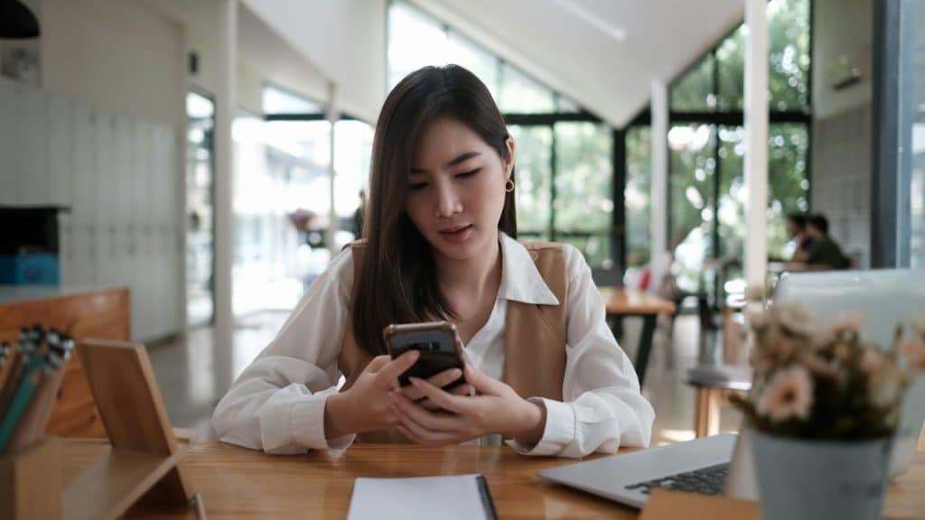 Young woman looking down intently at her mobile phone while sitting at a wooden desk.