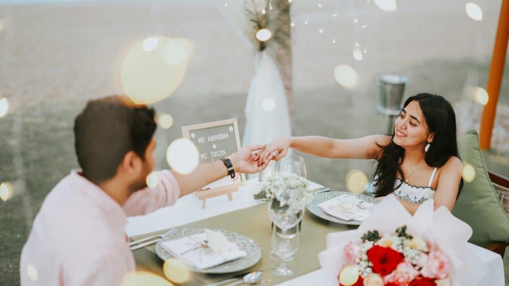 Couple holding hands across a romantic outdoor dinner table with a bouquet of flowers.