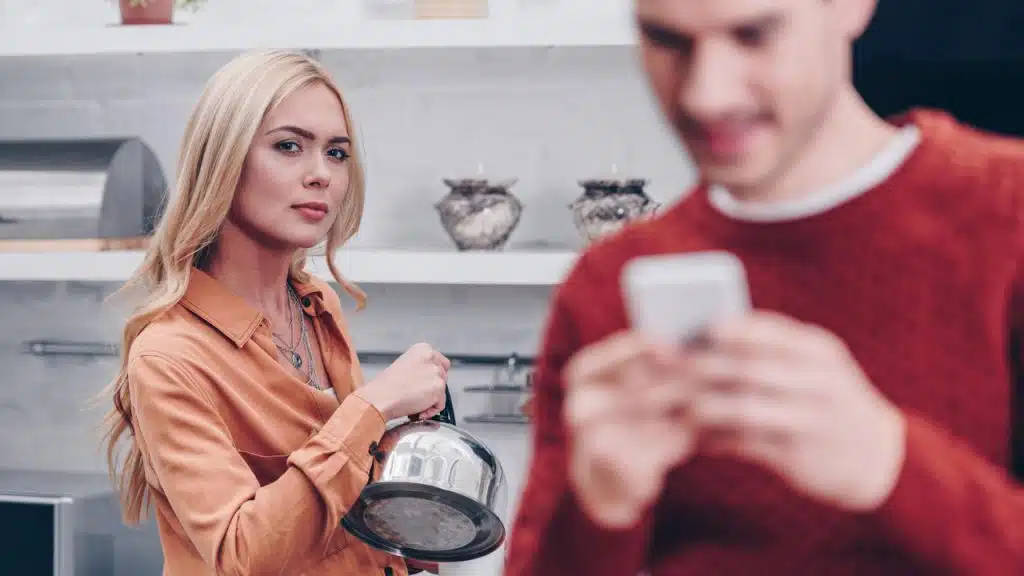 Woman holding a kettle and looking at the camera while a blurred man checks his phone.
