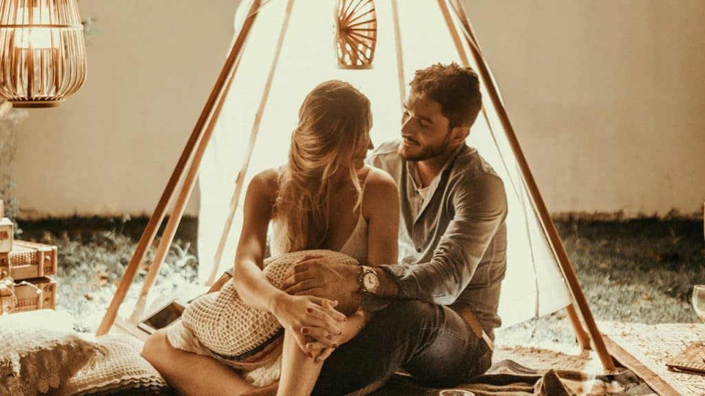 A man and woman sitting together on the ground under a makeshift tent in their living room. 