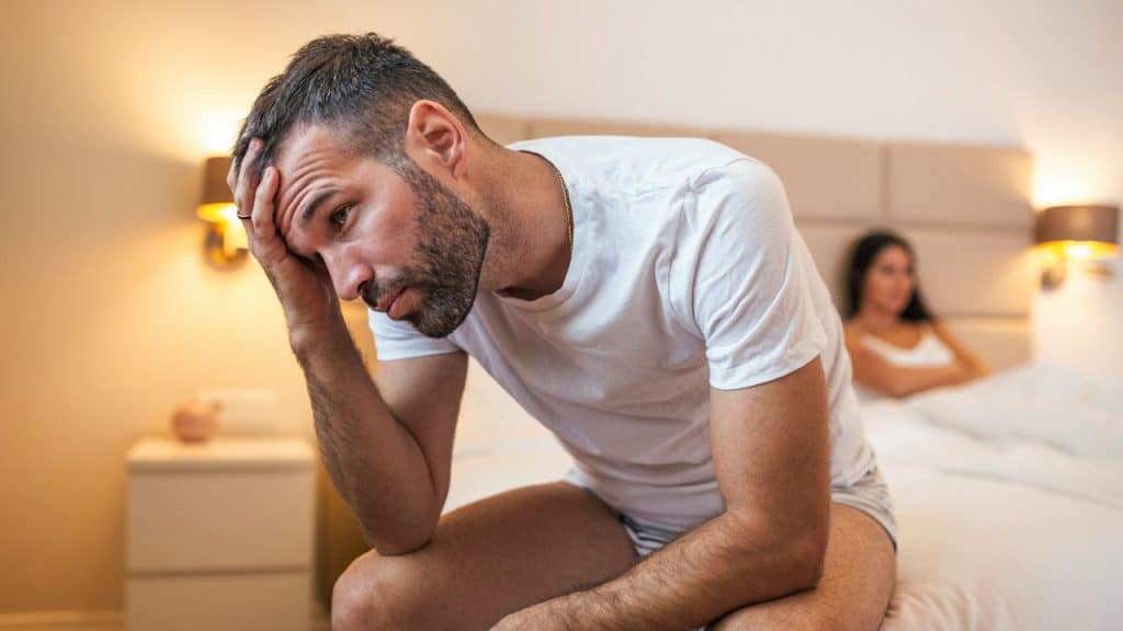 Man sitting on a bed with a distressed expression, hand on his forehead, woman in background.
