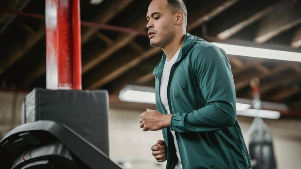 A man in a green jacket running on a treadmill in a gym.