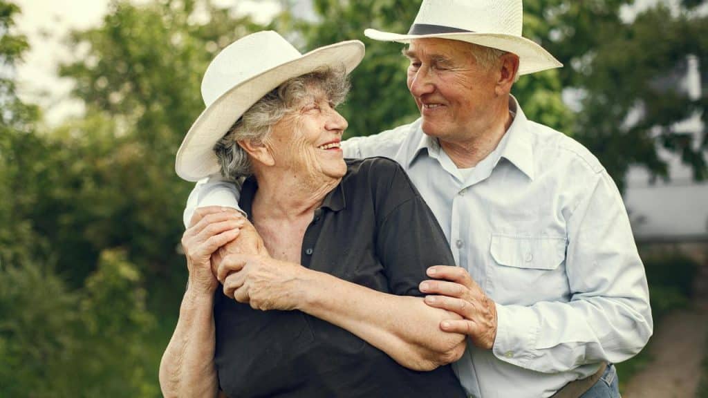 An elderly couple wearing white hats smiling and embracing outdoors.