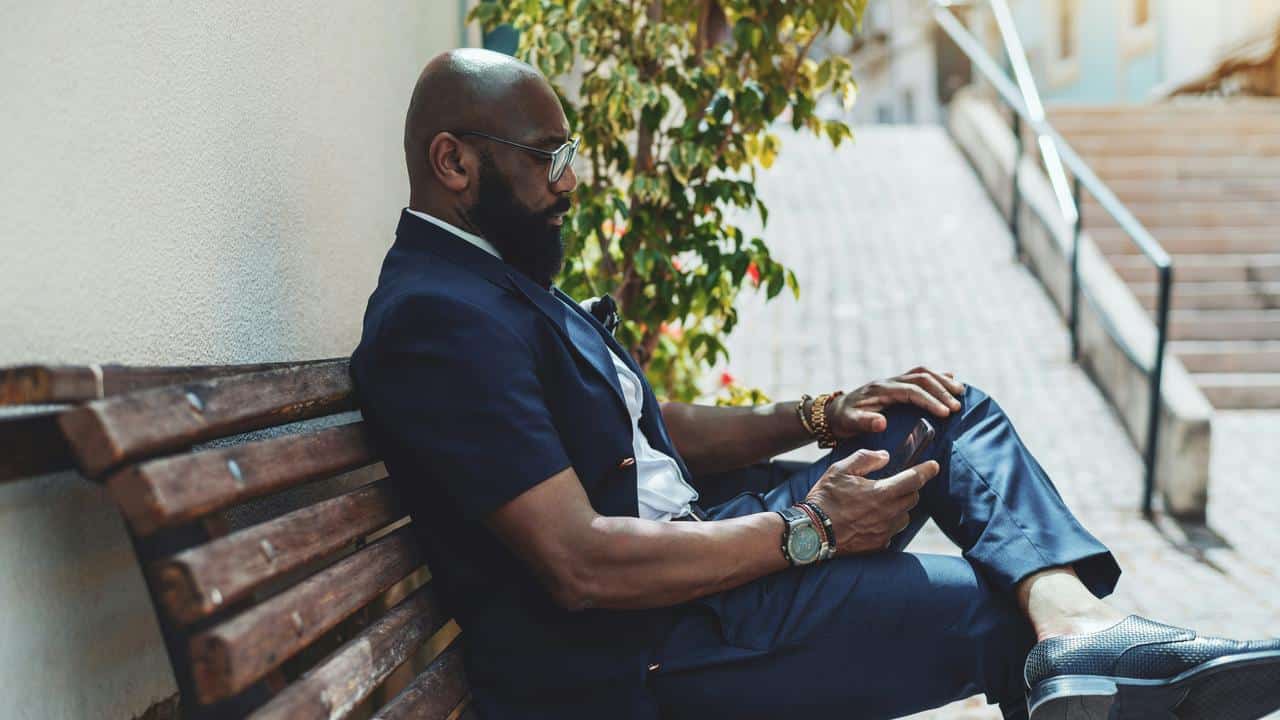 A man in a suit sitting on a bench outdoors while looking at his phone.