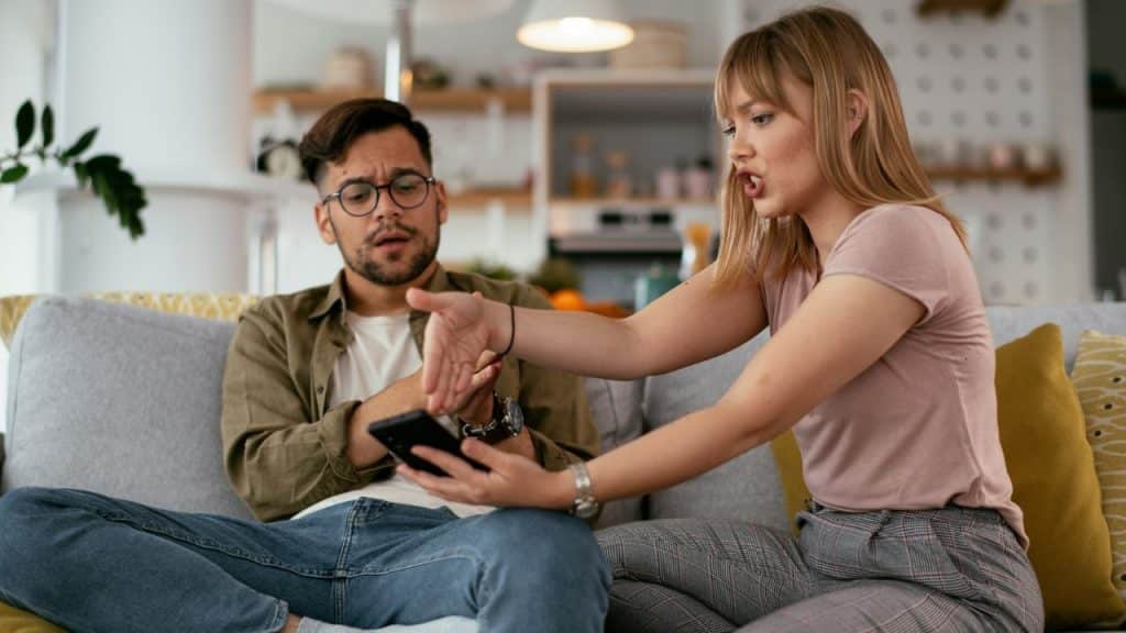 A woman argues with a man holding a phone while sitting on a living room couch.