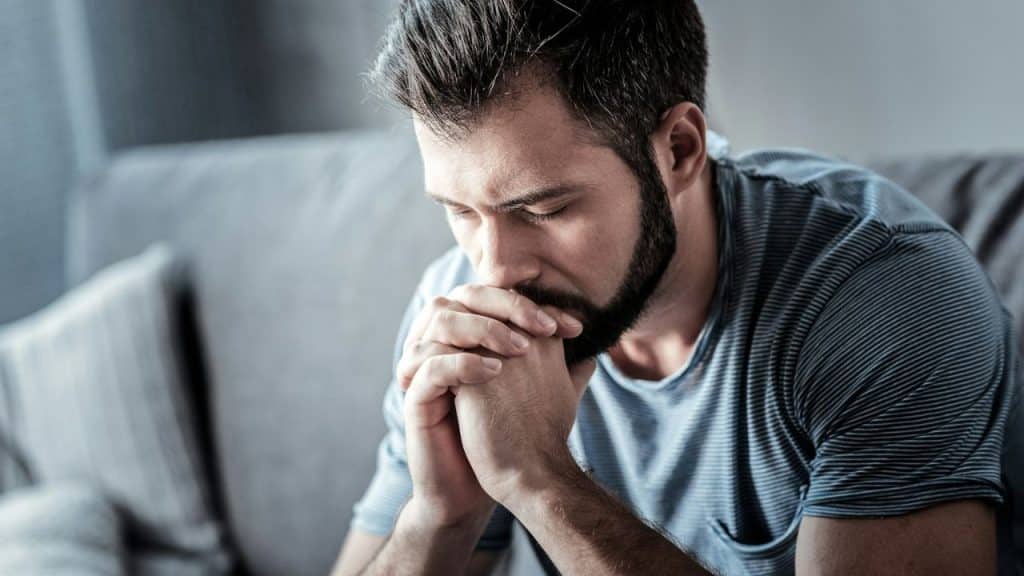 A bearded man sits with his hands clasped under his chin, looking down in distress.