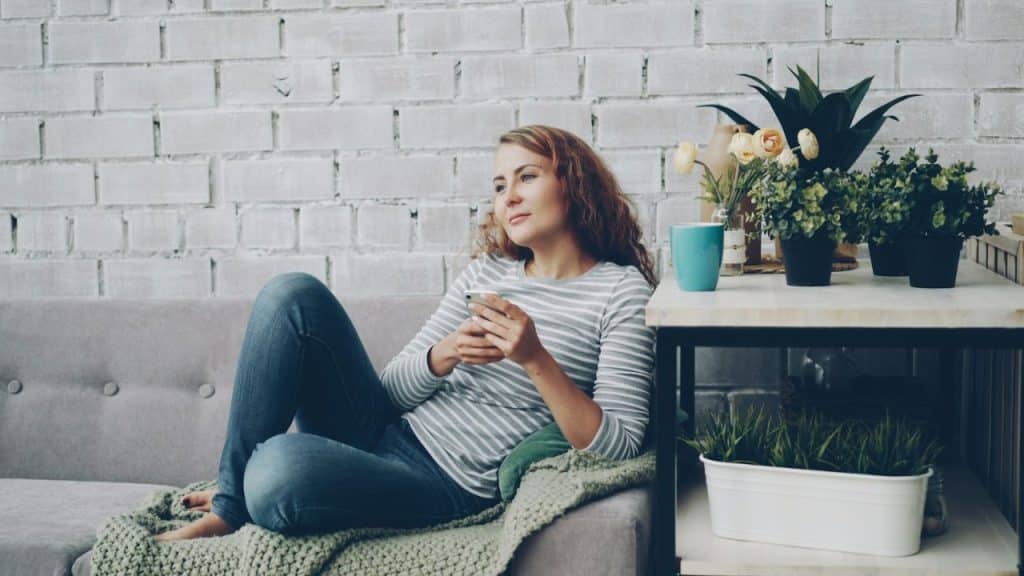 A woman sits comfortably on a couch, holding a phone, looking away from the camera.