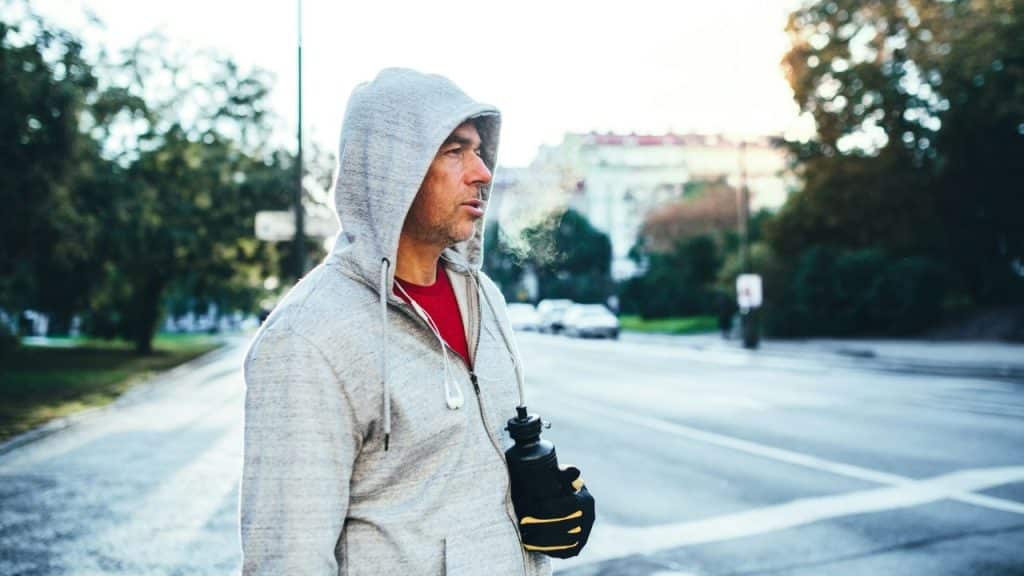 A man in a hooded sweatshirt stands outside, exhaling steam, holding a water bottle.
