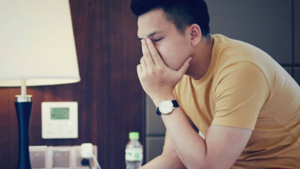 A man in a yellow shirt sits with his hand covering his eyes near a bedside lamp.