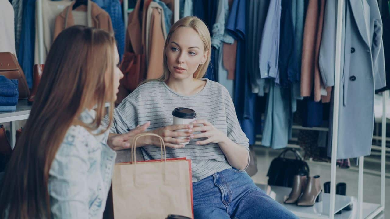 Two women talk in a clothing store, one holding a coffee cup and a shopping bag is visible.