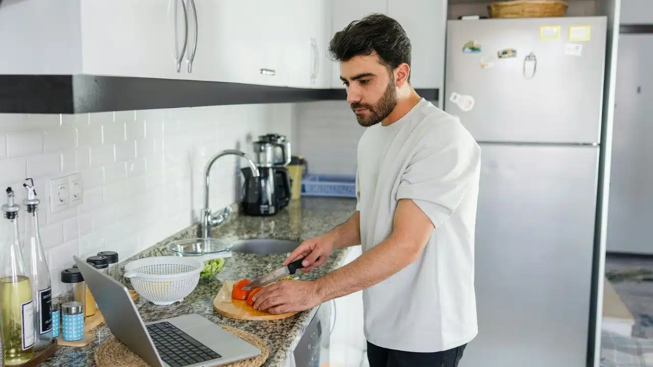 Man in a white shirt cutting a tomato on a wooden board in a modern kitchen.