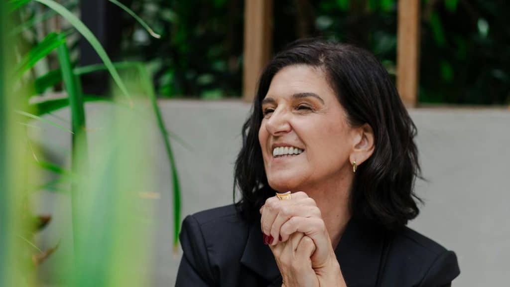 A smiling woman with short dark hair sits in a green, plant-filled setting.