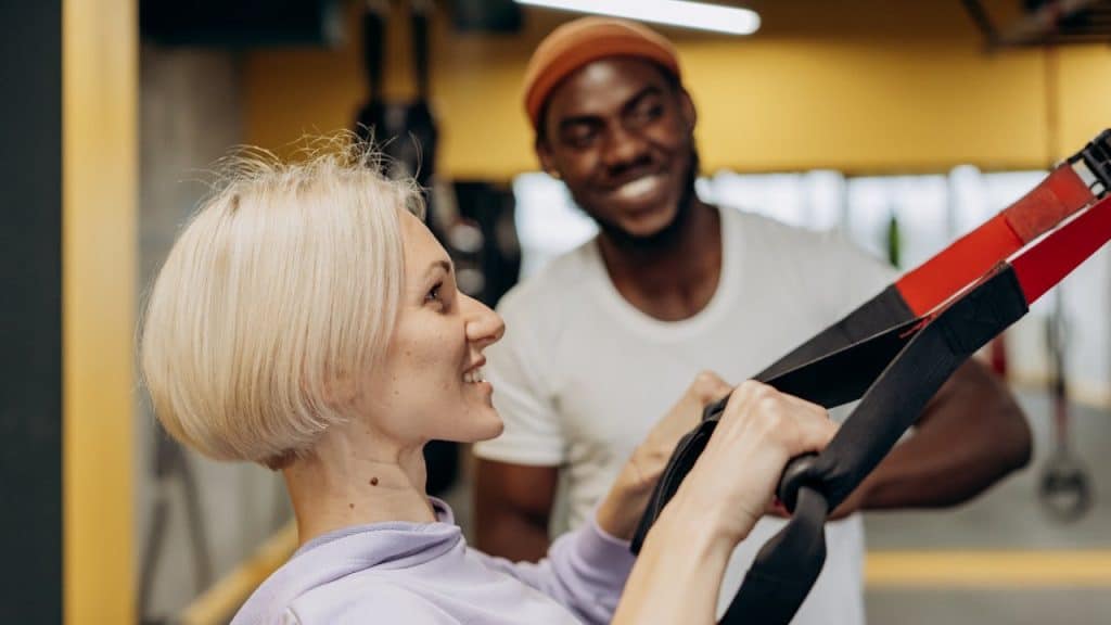 Smiling woman doing suspension training with a male trainer watching her.
