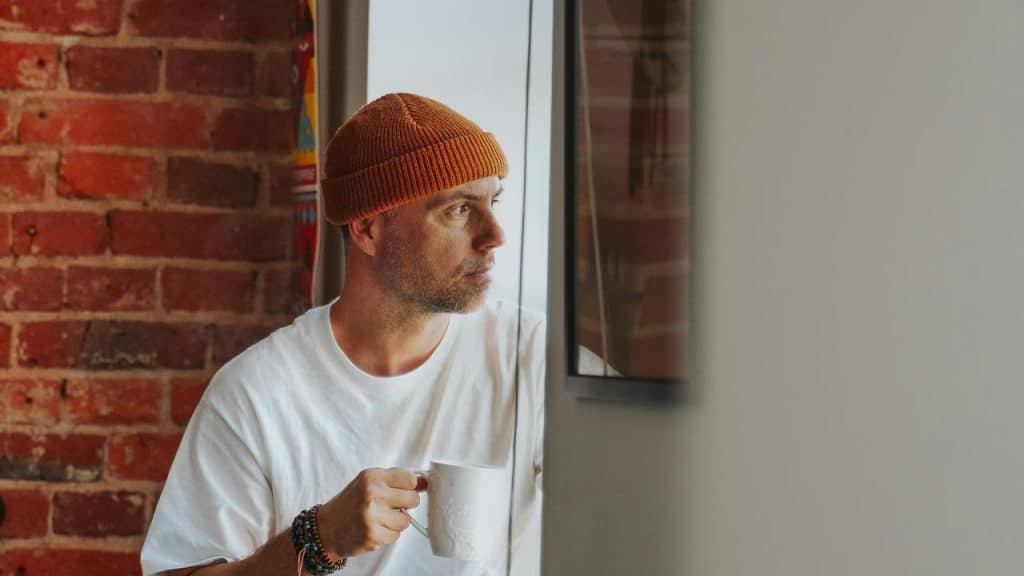 A man in a beanie holds a coffee mug while looking out a window thoughtfully.