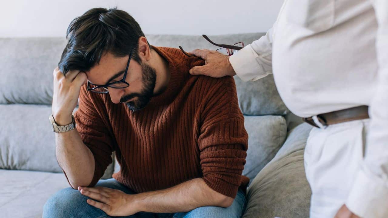 A person's hand rests on the shoulder of a sad, seated man with glasses and a beard.