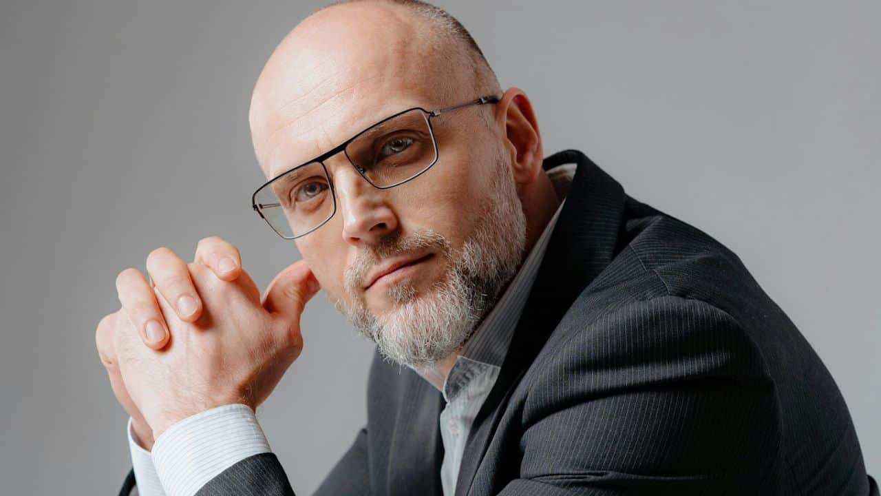 A bald man with a gray beard and glasses is looking intently at the camera.