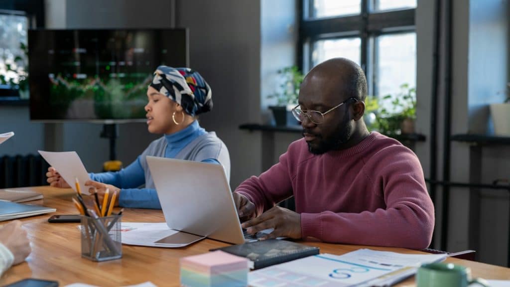 A man and a woman are sitting at a conference table, with the man working on a laptop and the woman reviewing papers.