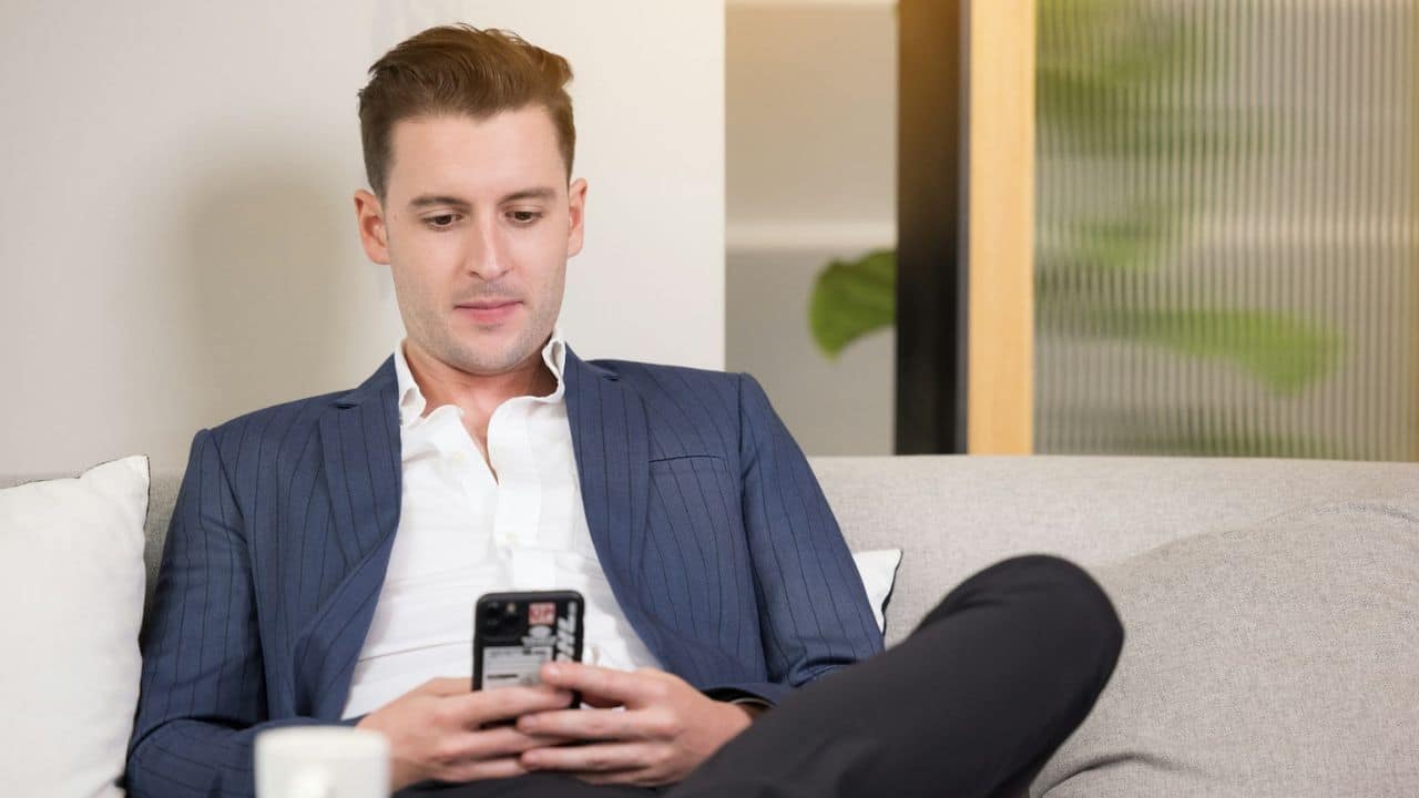 A man in a suit jacket is sitting on a sofa looking intently at his cell phone.