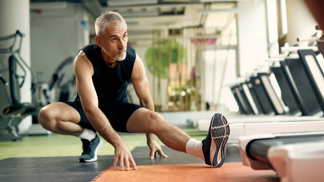 An older man with gray hair stretching his legs on a mat in a gym.